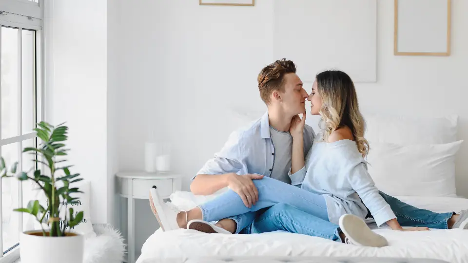 Par sidder tæt sammen på en hvid seng i et lyst soveværelse, et billede på sexdating for par